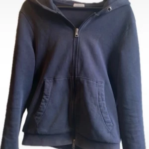 Mörkblå huvtröja från Moncler - Säljer en snygg mörkblå huvtröja från Moncler med dragkedja och praktiska fickor framtill. Tröjan har en broderad Moncler-logga på ärmen och en randig detalj på huvan. Perfekt för en stilren och bekväm look.