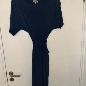 Mörkblå klänning från bubbleroom - Elegant mörkblå klänning  med knytband i midjan. En slit finns även på sidan. Klänningen har kortärmade ärmar och en lös passform som ger en avslappnad look. Perfekt för både vardag och festliga tillfällen. Använd en gång 