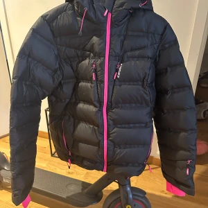 Svart dunjacka med rosa detaljer från Everest - Snygg svart dunjacka från Everest med rosa dragkedjor och foder. Jackan har en justerbar huva och långa ärmar med rosa muddar. Perfekt för kyliga dagar!