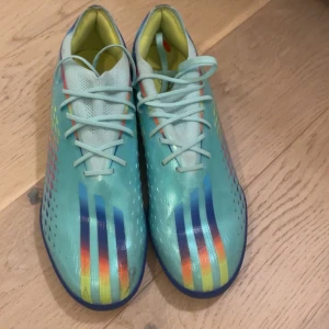 Turkosa fotbollsskor med regnbågsdetaljer - Snygga turkosa fotbollsskor med regnbågsfärgade detaljer och snörning. Perfekta för fotbollsplanen med en modern design och bekväm passform.