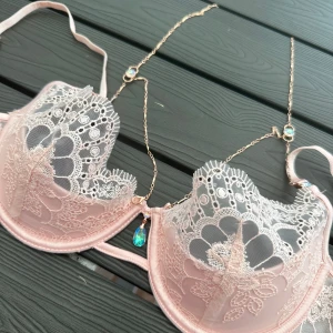 Rosa spets-bh med smyckesdetaljer - Elegant rosa bh i spets med vackra blommönster och smyckesdetaljer. Den har justerbara axelband och en dekorativ kedja med glittrande stenar. Aldrig använd med prislapp. Storlek 80B