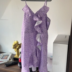 Lila blommig klänning med volang - Säljer en söt lila klänning med små vita blommor och volangdetalj framtill. Klänningen har smala axelband och är ärmlös, perfekt för varma dagar. Lätt och luftig modell som ger en somrig känsla.