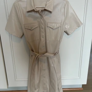 Beige klänning i skinnimitation och bälte - Snygg beige kortklänning i skinnimitation med korta ärmar, krage och knappar framtill. Två bröstfickor med lock och ett matchande bälte i midjan. Använd 1 gång, super cool från BikBok.