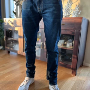 Mörkblå jeans från Nudie Jeans - Säljer ett par klassiska mörkblå jeans från Nudie Jeans med orangea sömmar och fem fickor. Jeansen har normal passform och raka ben, tillverkade i bomull. Perfekta för dig som gillar stilrena och tidlösa jeans. Modellen är tight terry 