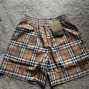 Rutigta Burberry shorts i beige - Snygga shorts från Burberry i klassiskt rutigt mönster med beige bas och inslag av svart, vitt och rött. Elastisk midja och lätt material, perfekta för sommaren. Ikonisk design som sticker ut.