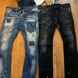 DSQUARED2 jeans med slitningar och färgstänk - Två par DSQUARED2 jeans, ett par blå och ett par svarta. Båda har slitna detaljer, färgstänk och trasiga partier för en edgy look. Klassisk femficksmodell med dragkedja och knappgylf. Märkesdetaljer på dragkedja och insida. 650 styck. Båda för 1100kr