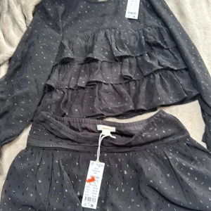 Svart blus med volanger och glitter detaljer, även matchande kjol - Supersöt svart blus och kjol från gina tricot. Med svarta glittriga prickar. Säljer då dom är för små och aldrig kommer till användning. Dom är aldrig använda, tröjan är i storlek 146/152 och kjolen är i 158/164💞