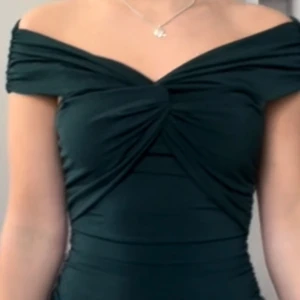 Mörkgrön offshoulder topp - Snygg mörkgrön topp med offshoulder-modell och twistad detalj framtill. Toppens material är stretchigt och sitter tight mot kroppen. Perfekt för dig som vill ha en elegant och trendig look.