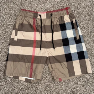 Rutiga shorts  från Burberry - Säljer ett par klassiska badshorts från Burberry med rutigt mönster i beige, svart, vitt och rött. Shortsen har snörning i midjan och sidofickor. Perfekta för sommaren och strandhäng.