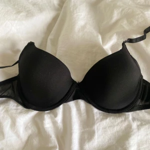 Svart BH från H&M - Svart BH från H&M med vadderade kupor och justerbara axelband. Klassisk modell med slät yta och hakknäppning bak. Perfekt för dig som vill ha en enkel och stilren BH i garderoben. Ena bandet är av men det går att antingen sy fast eller ta bort banden helt