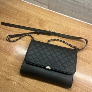 Svart quiltad axelväska från Don Donna - Snygg svart axelväska från Don Donna med quiltat mönster på framsidan och kedjedetaljer på axelremmen. Väskan har ett lock med metallknäppe och är tillverkad i syntetmaterial. Perfekt storlek för det viktigaste och stilren design. Lite små slitage på sidorna , ser bara om man tittar nära. 