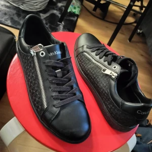 Svarta sneakers från Calvin Klein med dragkedja - Säljer ett par svarta Calvin Klein sneakers med snyggt mönstrad sida och silverfärgad dragkedja. Skorna har snörning, rund tå och platt sula. Perfekta för dig som gillar stilrena och moderna detaljer.