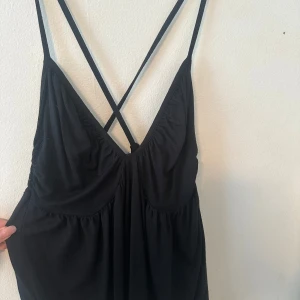 Svart linne med kors i ryggen - Säljer ett svart linne med smala korsade axelband i ryggen och djup v-ringning. Linnen har en lätt rynkad byst och resår i ryggen för extra komfort. Perfekt för varma dagar eller sommarens tillfällen.