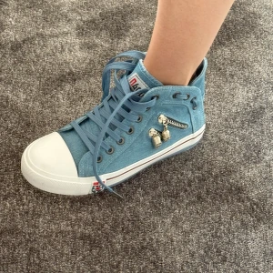 Blå höga sneakers med dödskalle-detaljer - Säljer ett par blå höga sneakers från LUODAN med vita gummisulor och snörning. Skorna har en liten dragkedja på sidan och coola dödskalle-berlocker som detalj. Perfekta för dig som vill sticka ut med din stil! Pris kan diskuteras