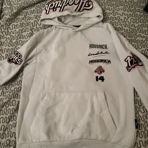hoodrich hoodie - Vit hoodie från Hoodrich med flera svarta och röda tryck på bröstet, ärmarna och huvan. Stor magficka framtill och huva med broderad logga. Perfekt för en streetwear-look.