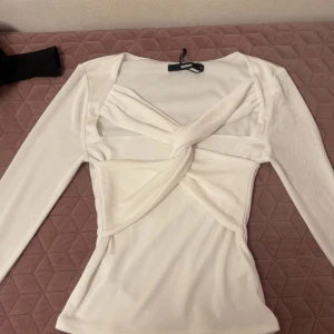 Vit långärmad topp med twistdetalj - Säljer en vit långärmad topp från BikBoK med unik twistdetalj framtill och figurnära passform. Toppen har en snygg skärning över bröstet och är tillverkad i ett mjukt material. Perfekt för att lyfta din outfit! I storlek S