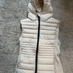 Vit dunväst med huva från Zara - Säljer en snygg vit dunväst från Zara med huva och svart dragkedja. Västen är quiltad och har en sportig look som passar perfekt till lager-på-lager-stil. Perfekt för kyliga dagar när du vill hålla värmen utan att kompromissa med stilen.