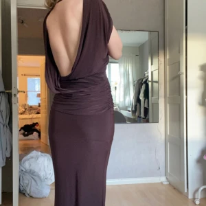 Mörkbrun långklänning med öppen rygg - Säljer en elegant mörkbrun långklänning med draperad effekt och djup öppen rygg. Klänningen har en åtsittande passform och är ärmlös med rund halsringning framtill. Det är Stretchigt material, passar xs-m. Aldrig använd förut och prislapp sitter kvar. Går att få billigare vid snabb affär. 