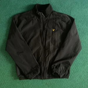 Svart vindjacka från Lyle & Scott - Obs!!! Barnstorlek. Säljer en svart vindjacka från Lyle & Scott med dragkedja och klassisk gul logga på bröstet. Jackan har hög krage, långa ärmar och elastiska muddar. Perfekt för blåsiga dagar och enkel att matcha med det mesta. Mycket fint skick, skulle nog säga aldrig använd!