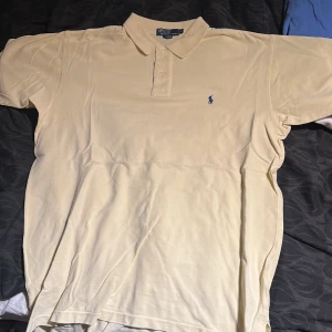 Gul piké t-shirt från Ralph Lauren - Snygg gul piké t-shirt från Ralph Lauren med klassisk krage och knappar framtill. Liten blå broderad logga på bröstet. Perfekt för en avslappnad men stilren look. Gamla taggen 