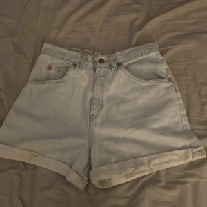 Ljusblå jeansshorts från Levi's - Säljer ett par vintage ljusblå jeansshorts från Levi's med hög midja och uppvikta benslut. Shortsen har fem fickor och synliga nitar framtill. Perfekta för sommaren och ger en avslappnad vintagekänsla. Storlek 7 = 36