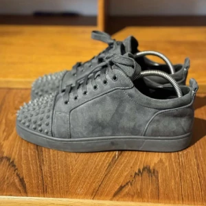 Gråa Louboutin  - Säljer ett par mörkgrå sneakers i mocka med coola nitar på tån. Skorna har snörning och en platt sula. Perfekta för dig som vill sticka ut med din stil. Självfallet är dem äkta. Endast seriösa köpare.