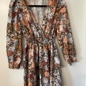 Blommig klänning med v-ringning - Superfin blommig klänning med djup v-ringning och långa ärmar. Klänningen har smock i midjan och volangdetaljer vid kjolen. Färgerna går i orange, blått, grönt och vitt. Perfekt för vår och sommar!