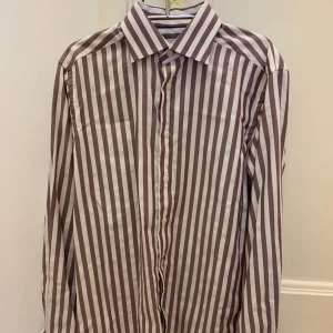 Brun- och vitrandig skjorta från Eton - Randig bomulls skjorta från Eton. Skjortan har lång ärm, knappar framtill och en stilren krage. Perfekt för dig som gillar tidlös och stilren design. Stoleken är medium. Fler bilder går att lösa! Nypris ca 1500kr