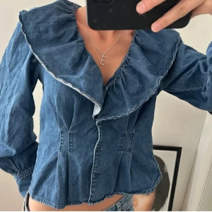 Jeans topp - Säljer en blå jeansskjorta med stor volangkrage och knappar framtill. Skjortan har lång ärm och figurnära passform, perfekt för dig som vill ha något unikt och trendigt i garderoben.