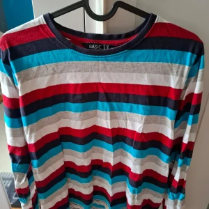 Randig långärmad tröja från BASIC U - Säljer en långärmad tröja från BASIC U med färgglada horisontella ränder i rött, blått, vitt, grått och mörkblått. Tröjan har en klassisk rund halsringning och är perfekt för dig som gillar en färgstark stil.