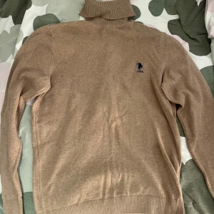Beige polotröja från U.S. Polo Assn. - Säljer en klassisk beige polotröja från U.S. Polo Assn. med broderad logga på bröstet. Tröjan har lång ärm och ribbade muddar vid ärmslut och nederkant. Perfekt för lager-på-lager och stilrena outfits.