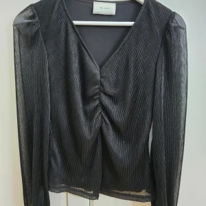 Svart plisserad topp från Neo Noir - Säljer en svart plisserad topp från Neo Noir med v-ringning och rynkdetalj framtill. Toppen har långa, lätt transparenta ärmar och en smickrande passform. Perfekt att styla till olika tillfällen!