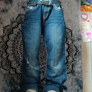 Blå jeans från Hydraulic med detaljer - Säljer ett par blå jeans från Hydraulic med slitningar och dekorativa sömmar samt nitar på bakfickan. Jeansen har normal passform och klassisk femficksdesign. Perfekta för dig som gillar lite extra detaljer på dina jeans.