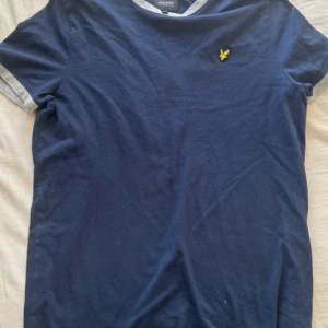 Blå t-shirt från Lyle & Scott  - Mörkblå t-shirt från Lyle & Scott Junior med gråa detaljer vid hals och ärmslut. Klassisk passform och liten gul logga broderad på bröstet. I jätte bra skick och inte använd så mycket 