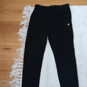 Svarta mjukisbyxor från Lyle & Scott - Svarta mjukisbyxor från Lyle & Scott med gul broderad logga på framsidan. Byxorna har elastisk midja och en bakficka. Perfekta för en avslappnad stil. Aldrig använda