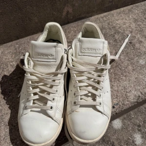 Vita Adidas Stan Smith sneakers - Säljer ett par klassiska vita Adidas Stan Smith sneakers med diskret perforerat mönster på sidorna och vit mockadetalj vid hälen. Skorna har snörning och är tillverkade i skinn. Perfekta för en clean och stilren look. Observera att skorna har lite slitningar som man ser på bilderna🩷