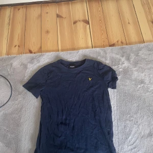 Mörkblå t-shirt från Lyle & Scott Junior - Säljer en mörkblå t-shirt från Lyle & Scott Junior med klassisk passform och korta ärmar. T-shirten har en liten gul broderad logga på bröstet och är tillverkad i mjuk bomull. Perfekt för en avslappnad stil.