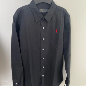 Svart skjorta från Polo Ralph Lauren - Klassisk svart skjorta från Polo Ralph Lauren med vit knappstängning och rött broderat logotyp på bröstet. Skjortan har lång ärm och en stilren, tidlös design som passar till många tillfällen.