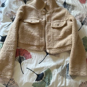 Beige teddyjacka från SHEIN - Mysig beige teddyjacka från SHEIN med croppad passform. Jackan har två bröstfickor med lock, knappar framtill och lång ärm. Perfekt för kyliga dagar när du vill vara både varm och trendig.