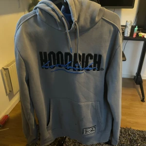 Grå hoodie från Hoodrich - Säljer en grå hoodie från Hoodrich med svart och blå logga framtill. Tröjan har huva med dragsko, magficka och ribbade muddar. Perfekt för en avslappnad stil.