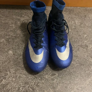 Blå Nike CR7 fotbollsskor med högt skaft - Säljer ett par blå Nike CR7 fotbollsskor med högt strumpskaft och stora vita swoosh-loggor. Skorna har snörning och är designade för bra grepp på planen. Perfekta för dig som vill sticka ut på fotbollsplanen.