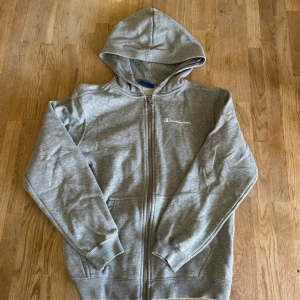 Grå hoodie från Champion - Grå hoodie från Champion med dragkedja framtill och huva. Tröjan har långa ärmar och två fickor på sidorna. Champion-loggan är broderad på bröstet. Perfekt för en avslappnad stil.