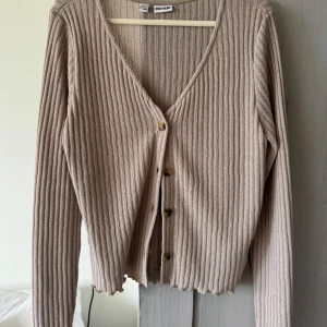 Beige ribbad kofta från Bodyflirt - Säljer en beige ribbad kofta från Bodyflirt med v-ringning och knappar framtill. Koftan har långa ärmar och en croppad, vågig nederkant. 