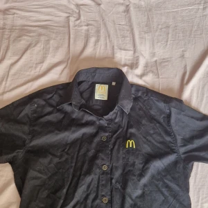 Svart McDonald's skjorta - Svart skjorta från McDonald's Professional Workwear Edition med klassisk krage och knäppning framtill. Broderad gul McDonald's-logga på bröstet. Perfekt för dig som vill ha en unik och ikonisk skjorta.