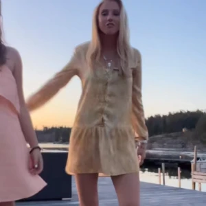 Gul långärmad kortklänning med knappar - Säljer en söt gul kortklänning med långa ärmar och knappar framtill. Klänningen har en lätt volang nertill och är perfekt för varma dagar. Den har en mjuk, lite glansig yta och en avslappnad passform.