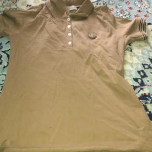 Beige pikétröja från Moncler - Säljer en beige pikétröja från Moncler med klassisk krage och knappslå med fem vita knappar. Tröjan har korta ärmar och en broderad Moncler-logga på bröstet. Perfekt för en stilren och avslappnad look.