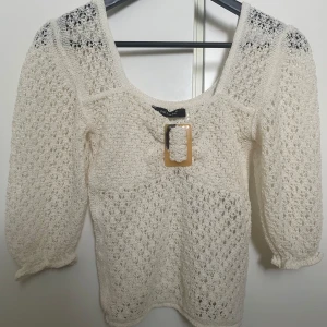 Vit virkad blus med spänne - Säljer en söt vit virkad blus med trekvartsärmar och ett dekorativt spänne framtill. Blusen har en djup urringning och är tillverkad i ett luftigt material som passar perfekt till våren och sommaren.