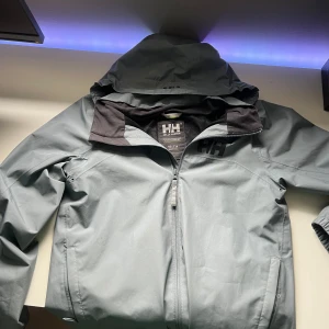 Ljusgrå vindjacka från Helly Hansen - Säljer en ljusgrå vindjacka från Helly Hansen med huva och dragkedja framtill. Jackan har en stilren design med HH-logga på bröstet och är perfekt för blåsiga dagar. Passar dig som gillar funktionella och snygga ytterplagg. Den här målarfärg på två ställen vilket finns på bilderna därför har den så lågt pris. Skriv om ni har några frågor