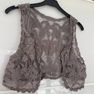 Grå spetsväst med blommigt mönster - Säljer en grå väst i spets med vackert blommigt mönster. Västen är kort och har en öppen front, perfekt för att lägga till en romantisk touch till din outfit.