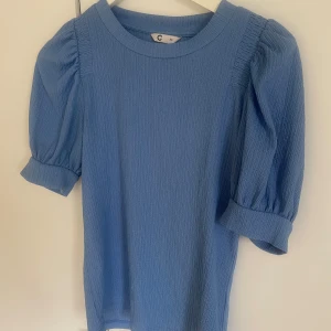 Blå blus med puffärm från Cubus - Säljer en ljusblå blus från Cubus med puffärmar och rund halsringning. Klänningen har en strukturerad yta och trekvartsärm med lätt volym. Perfekt för dig som gillar en feminin och stilren look.